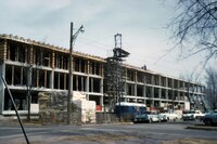 Multi-story building under construction along a city street, with scaffolding, construction materials, utility poles, and parked cars visible.
