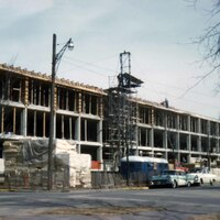 Multi-story building under construction along a city street, with scaffolding, construction materials, utility poles, and parked cars visible.